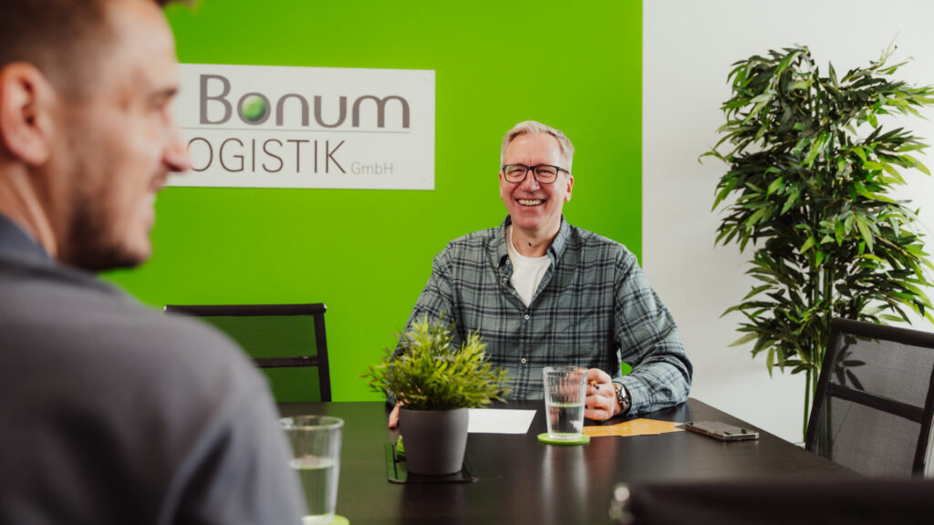 Zwei Personen im Besprechungsraum von Bonum Logistik, eine sitzt vor grüner Wand mit Bonum‑Logo, auf dem Tisch stehen Wasser und Pflanzen